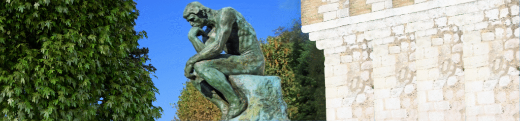 A photo featuring Auguste Rodin's sculpture The Thinker positioned outdoors against a backdrop of green trees and blue sky. On the left side, there is a dense leafy tree, while on the right, a classical stone building with columns is partially visible.