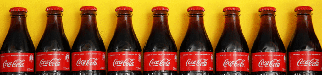 A row of classic Coca-Cola glass bottles with red labels and caps, placed against a bright yellow background.