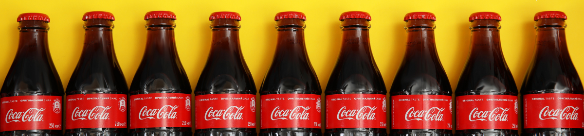 A row of classic Coca-Cola glass bottles with red labels and caps, placed against a bright yellow background.