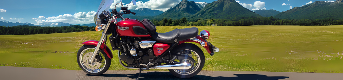 A red Triumph motorcycle parked on a scenic road with a backdrop of green fields, forest, and majestic mountain ranges under a bright blue sky with scattered clouds.