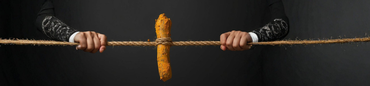 Two hands pull a rope taut from opposite sides against a dark background, with a turmeric coated root tied at the center.