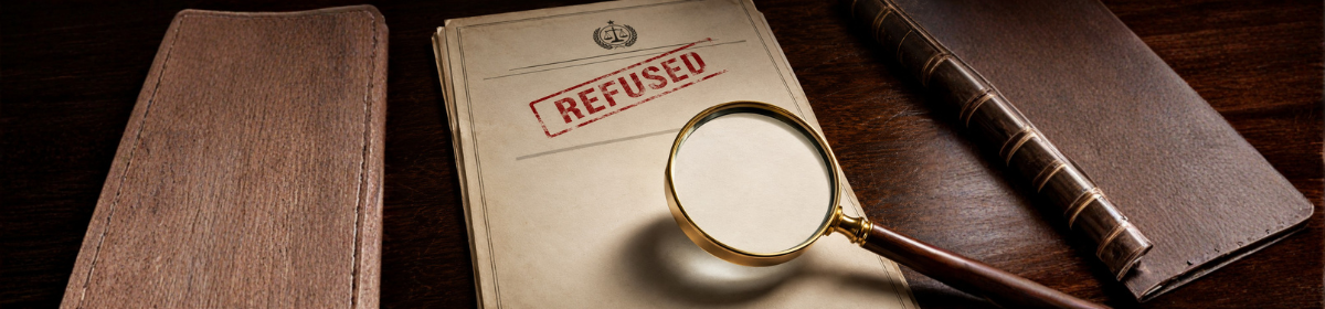 Patent refusal document stamped “Refused” with a magnifying glass on a wooden desk, representing legal review of a rejected patent application.