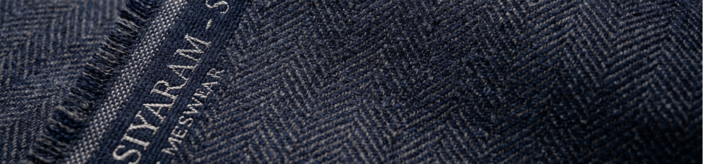 Close-up macro photograph of premium suiting fabric in deep navy and charcoal grey, with a fine herringbone or pinstripe weave pattern. Along the selvedge edge, the word "Siyaram" appears subtly woven or embossed into the cloth in a slightly lighter thread.