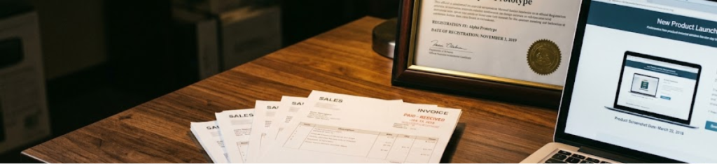 A close-up, dimly lit still-life on a polished wooden table featuring a stack of sales invoices and a laptop displaying a product webpage. Beside these items is a framed design registration certificate. The scene is illuminated by a warm amber spotlight, visually representing a courtroom-themed concept where a plaintiff's own early-dated documents serve as key evidence of prior publication against their later-dated registration